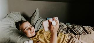 Ung, smilende jente ser på mobiltelefon liggende i senga.